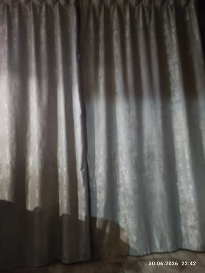 This is a close-up, horizontally oriented image of grey, pleated curtains. The fabric has a textured, slightly shimmering appearance due to the way light reflects off its folds. There are multiple horizontal pleats creating a rhythmic pattern of light and shadow across the surface. The top and bottom sections of the image show the curtains in slightly different light conditions, with the top appearing darker and more shadowed, while the bottom is brighter.

To the right side of the image, there is text overlaid, which appears to be a date and time stamp: "30.06.2026 22:42". This indicates the image was taken on June 30, 2026, at 10:42 PM. The presence of this timestamp and the indoor setting (suggested by the curtains) imply the image was taken during the evening. The lighting in the photograph is dim, contributing to the dark shadows within the folds of the curtains. There is a hint of a darker, possibly wooden surface on the far right edge of the frame. No people or specific objects are clearly discernible, the focus is entirely on the texture and form of the curtains. The location context provided suggests Iskitim, Russia.