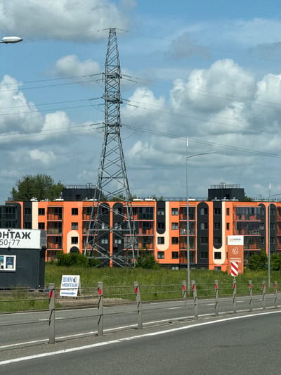 The image captures a street view in Oktyabrsky, Russia, dominated by a tall, metallic electricity pylon situated in the foreground. Behind the pylon, a modern, multi-story apartment building with an orange and black facade stands out. The building features numerous windows and balconies, some of which are enclosed. In front of the apartment building, a patch of green grass and some trees are visible. To the left of the pylon, a dark, rectangular structure with a white sign that reads "МОНТАЖ" and a phone number is partially visible. Further to the right, another sign with a chicken logo and text that appears to be "КОМОС" is displayed on the apartment building. The sky is partly cloudy with a bright blue sky and white cumulus clouds. A street lamp with a light fixture is positioned to the right of the pylon. A road with white lane markings is in the lower part of the image, with a metal barrier separating it from the grassy area. There are no people visible in the scene.