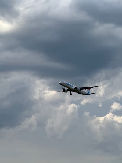 A commercial airliner, identified by the "Luxair" logo on its fuselage and the flag of Luxembourg on its tail, is captured mid-flight against a dramatic, overcast sky. The airplane, a twin-engine jet, is angled slightly upwards, suggesting it is either taking off or climbing after landing. Its landing gear is extended. The surrounding environment is characterized by heavy, dark gray clouds with some lighter patches indicating the presence of sunlight breaking through. The photograph appears to have been taken from below the aircraft, looking up. The setting is implied to be near Hahn, Germany, likely at or around Frankfurt-Hahn Airport, a common operational area for Luxair. The image is devoid of any people or other discernible objects in the foreground or background, focusing solely on the aircraft and the sky.