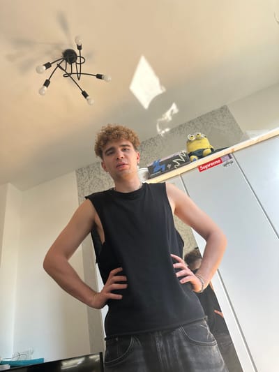 A young man with curly blonde hair stands in a room, his hands on his hips and his gaze directed slightly upwards and to the left. He is wearing a black sleeveless shirt and dark denim jeans. Behind him, on a shelf, a Minion toy and several boxes are visible. A modern chandelier hangs from the ceiling above him, casting shadows on the wall. The room appears to be illuminated by natural light, possibly from a window. The overall impression is a casual, personal space. No specific location in Hamm, Germany, can be determined from the visual information provided.