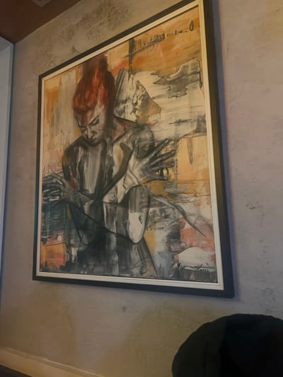 A vertically oriented, rectangular framed painting is affixed to a textured light grey wall within a building in Smederevska Palanka Municipality, Serbia. The artwork is an abstract-figurative piece depicting a human figure, likely female, rendered predominantly in shades of grey, black, and white, with prominent red hair styled in an updo. The figure's head is tilted downward, and both hands are dynamically positioned; the left hand is raised near the figure's chest or face, while the right hand is extended to the right, fingers splayed. The background of the painting features broad, abstract brushstrokes in warm tones of orange, beige, and darker grey, suggesting a dynamic, layered composition. A discernible artist's signature and the year "2022" are visible in the lower right corner of the painting. The artwork is enclosed within a frame featuring a thick dark outer border and a thinner white inner border. The wall itself exhibits a rough, stucco-like texture. In the upper left corner of the scene, a brown architectural element, possibly part of a ceiling or beam, is partially visible. The lower right foreground includes a partial view of a dark, soft object, consistent with a cushion or furniture armrest.