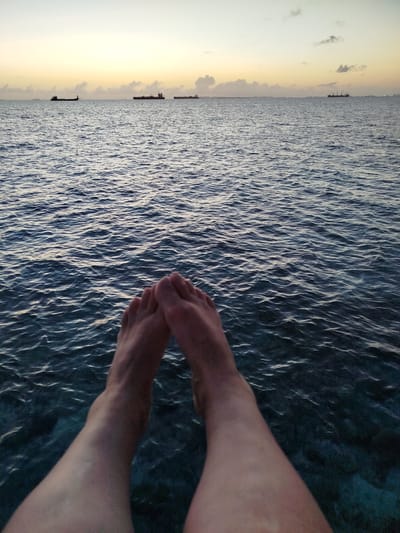 A first-person perspective shot shows two bare feet and legs dangling over a body of water, with small waves rippling on the surface. In the distance, several cargo ships are visible on the horizon against a pale orange and yellow sky, suggesting either dawn or dusk. The scene appears to be located in the ocean near the city of Gulhi Falhu, Maldives.