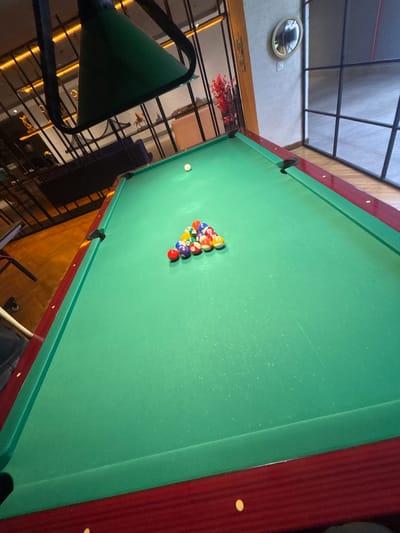 A pool table with a green felt surface and a burgundy frame is the primary subject of the image. A rack of billiard balls, including solid colors and stripes, is arranged on the table, with a white cue ball positioned separately. The pool table is situated in what appears to be an indoor recreational space, possibly a game room or bar. The background suggests a modern interior design with metal grilles, decorative lighting, and some ambient furniture. The location is identified as Kâğıthane, Turkey, implying this scene takes place within that district. There are no people visible in the frame.
