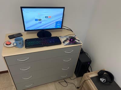 This image depicts an indoor workspace or gaming setup located in a corner of a room in Wegnez, Belgium. The central object is a light wood-topped, four-drawer grey dresser serving as a desk.

On the dresser top, a black Samsung computer monitor is illuminated, displaying the Opera web browser start page. To the left of the monitor, a black smartphone rests near a light blue mug with a cartoon character design and a small white plate holding a pink-frosted cupcake. Directly in front of the monitor is a black mechanical gaming keyboard with multi-colored RGB backlighting. To the right of the keyboard, a black gaming mouse with pink RGB lighting sits on a mousepad featuring a dark-robed figure.

Below and to the left of the monitor, a black desktop computer tower is situated on the floor, accompanied by a tangle of black cables connecting the various components. To the right of the computer tower, a small, light wood-grained end table with a dark surface holds a pair of black gaming headphones with white logo accents.

The room features plain, light-colored walls and a light-toned tiled floor with a brown baseboard. No individuals are present, and no active human interactions or actions are depicted.