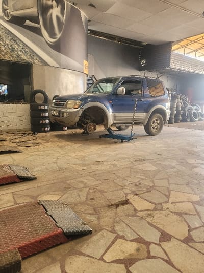 The image displays an interior view of an automotive service bay located in the Amaliada Municipal Unit, Greece. A two-door, blue over tan/silver Mitsubishi Pajero (or similar SUV) is centrally positioned, elevated by a blue hydraulic floor jack supporting its front left side. The front left wheel is absent, revealing the brake rotor and caliper assembly. The vehicle's three remaining wheels are on the ground. A long, slender metal rod leans against the vehicle's body, forward of the driver's door. No personnel are visible.

To the left of the vehicle, a stack of four tires, one mounted on a rim, rests against a low white brick wall. Behind this, a large cylindrical column is present, adorned with a graphic depicting a car wheel on a road. To the right, numerous stacks of tires are arranged along a dark-walled section of the facility. In the foreground, two red metal ramps with diamond plate surfaces are situated on the floor. The floor consists of irregularly shaped, light-colored flagstones set in reddish mortar, showing signs of wear and staining. The overhead structure features an industrial-style ceiling with exposed elements and a translucent roof section providing natural illumination.