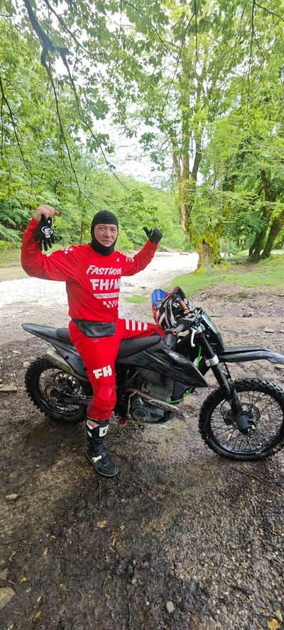 A person in red motocross gear, including a red jersey with "FASTHOUSE FHM" on it, red pants with white stripes and "FH" on the lower leg, black motocross boots, and a black balaclava, stands next to a black dirt bike in a wooded area. The person is holding a pair of black gloves in one hand and giving a thumbs-up with the other. The dirt bike has a helmet resting on the handlebars. The ground is muddy, and trees surround the scene. A small stream or clearing is visible in the background. The location is consistent with a rural area near Galitsyno, Russia, considering the type of vegetation and terrain.