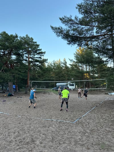 In Ozerki (Krasnodolinskaya), Russia, a group of six individuals is engaged in a game of volleyball on a sandy court surrounded by tall pine trees. The court is marked with white tape, and a white volleyball net is set up. The sky above is clear and blue.
On one side of the net, three people are playing. A person in a blue tank top and camouflage shorts is positioned near the net, facing away from the camera, appearing to be in motion. Another person, to their left, is wearing a blue shirt and shorts and appears to be watching the game. A third person, further back, is also dressed casually.
On the other side of the net, three individuals are actively involved in the game. One person in a bright yellow t-shirt and patterned shorts is in the foreground, facing the net. To their right, a person in a pink top and leggings is jumping, seemingly to hit the ball. Another person, further back, is wearing a dark shirt and shorts and is also in a dynamic pose, likely in the process of playing. A volleyball is visible in the air above the net.
In the background, several vehicles are parked, including a white camper van and a dark car. Tents and camping equipment are also present, suggesting a recreational setting. The ground is covered with sand, and scattered pine needles are visible. The overall atmosphere is casual and recreational.
