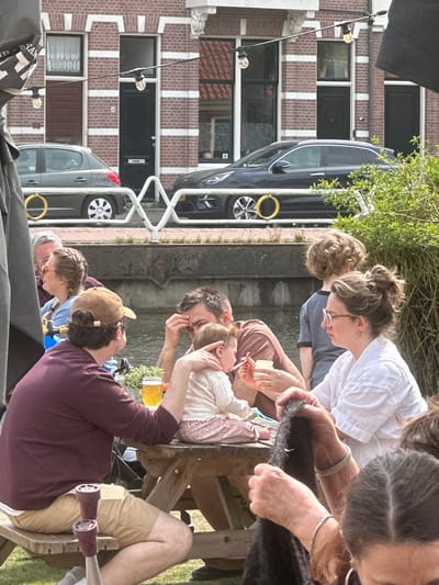 In the city of Utrecht, Netherlands, a group of people is gathered along a canal. A red brick building with white vertical accents and dark windows is visible in the background. A black car and a gray van, both with yellow circular accents, are parked adjacent to the building.
The group of people sits or stands near the canal. A baby is being held by an adult, and other individuals are interacting with or looking at the baby. An individual in a burgundy shirt and khaki shorts sits on a wooden chair, with a beer mug visible near them. There are several other adults standing or sitting around.
