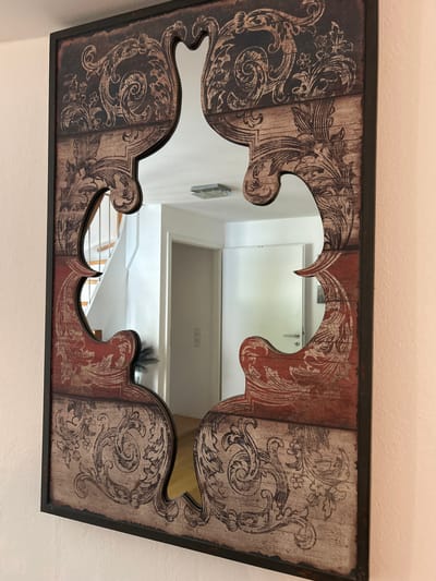 The image shows an ornate, dark-framed mirror with a decorative, cut-out design. The mirror's frame is adorned with a distressed wood-like pattern featuring scrolling botanical motifs in a muted red and beige color palette. The mirror itself reflects a hallway with a white door, a doorknob, a light fixture on the ceiling, and a staircase with wooden steps and a metal railing, suggesting an interior residential setting. A dark, feathery object is visible in the lower right reflection. The mirror is mounted on a light-colored, textured wall. The scene is static and appears to be a still photograph of the decorative mirror. There is no indication of the city of Velbert, Germany within the image itself.