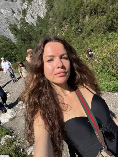 In Chemal, Russia, a woman with long, wavy brown hair takes a selfie while standing on a gravel path in front of a steep, rocky mountainside covered with green trees and shrubs. She is wearing a black strapless top with a red and green striped strap. A handbag with a patterned design is slung over her shoulder, and a pair of dark sunglasses hangs from the strap. In the background, a woman in a white shirt and grey pants walks away from the camera, and a child in a light green t-shirt is visible further down the path. Another person, wearing a dark t-shirt, is seen walking on the grassy hillside in the distance. The sun is casting shadows, indicating it is daytime.