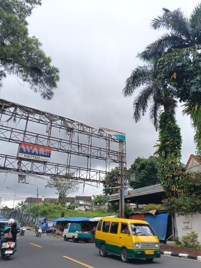 This image depicts a bustling street scene in Palayan, Philippines, under an overcast sky. The primary subjects are a road, various vehicles, a large dilapidated billboard, and dense tropical foliage.
In the foreground, an asphalt road occupies the lower portion of the frame, marked with a yellow solid line on its right edge. A yellow and green passenger van, possibly a public utility vehicle, is in motion towards the viewer on the right side of the road, its front blurred due to movement. On the left side of the road, a white scooter with a rider wearing a green jacket and white helmet is moving away from the viewer. Further back on the left, another motorcycle carries two individuals, one wearing a light blue helmet. Additional vehicles, including a small light blue pickup truck with a cargo bed, are visible parked or slowly moving on the roadside.
Dominating the left upper midground is a large, weathered metal billboard frame with tattered remnants of past advertisements. One section clearly displays a red and blue logo with text "NAKASHI" or "NAMASH," a phone number "53677322," and "MUNDING" below. Beneath the billboard, a raised embankment covered in green vegetation runs parallel to the road. Along the left edge of the road, several makeshift vendor stalls, covered with blue tarpaulins, are situated. Houses with various roof types are visible on the elevated terrain behind the stalls.
On the right side of the image, two tall palm trees with dense green fronds extend into the sky. The trunk of the closer palm tree is heavily covered in vines or epiphytes. Beneath these trees, abundant green foliage and a building with a reddish-brown roof and a light-colored facade are partially visible. A utility pole equipped with several lantern-style streetlights stands to the right of the billboard structure. A low, striped black and white barrier or curb runs along the far right edge of the road. Overhead utility lines are visible across the sky.