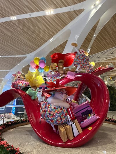 The image shows a whimsical art installation within an interior space, potentially an airport terminal, given the presence of structural beams and distant signage. The installation features oversized figures and objects arranged in a large, red, swirling structure. The figures are cartoonish, with colorful floral patterns and exaggerated features. One figure wears an orange hat and glasses. Accessories like shopping bags, a purple suitcase, a polka-dotted umbrella, and balloons are part of the display. The terminal's interior features wooden panels. Based on the location information, this is likely located in the Baiyun District, China.