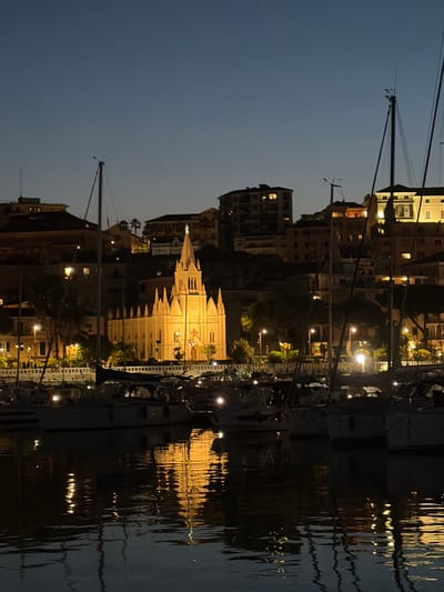 The image depicts a waterfront scene at dusk in the city of Imperia, Italy. Dominating the skyline is the illuminated Church of San Maurizio, a gothic revival style church with a prominent spire, glowing with a warm yellow light. It is situated on a hillside overlooking a harbor filled with sailboats. The water reflects the lights of the church and the surrounding buildings, creating shimmering patterns. Several buildings with illuminated windows are visible behind the church and along the waterfront, suggesting a residential or commercial area. The sky is a deep twilight blue, with no visible stars. The overall atmosphere is serene and peaceful.