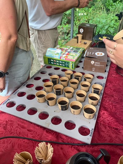 The image depicts an outdoor tasting event with small cups of dark liquid, presumably coffee, arranged in a grey tiered display. The display board has handwritten labels in black marker indicating different flavors or varieties. In the background, a table is covered with a red patterned cloth, on which boxes of coffee capsules and other items are placed. Two individuals are partially visible, one in the foreground with their arm and shoulder showing, wearing a light-colored tank top and denim shorts, and another person in the background wearing a white pleated shirt and beige pants. The setting appears to be a garden or outdoor space with lush green foliage in the background. The event is likely taking place in Hasselt, Netherlands, as suggested by the presence of a sign with "Hasselt" and other Dutch words.