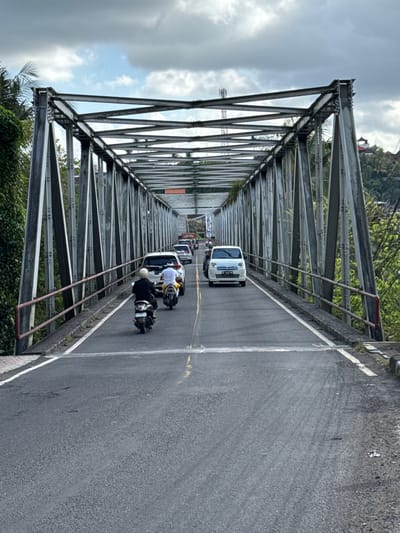 This is a vertical, eye-level, medium shot of traffic on a metal truss bridge in Klungkung, Indonesia. The bridge spans over a vegetated area and is made of grey metal girders and supports. The road surface is asphalt, marked with white lines.

In the foreground, a person on a scooter is riding away from the camera in the right lane. Behind them, a white SUV is also moving away. Further up the road, another scooter is visible, followed by a white van. In the distance, several other vehicles are seen on the bridge, including cars and motorcycles. The sky is overcast with grey clouds. Lush green vegetation is visible on both sides of the bridge, with some palm trees and hills in the background. The scene captures a moment of everyday traffic flow on a typical day.