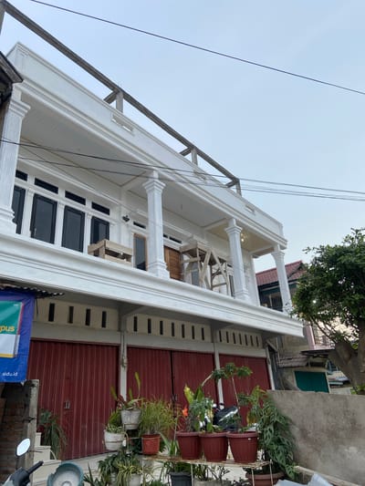 The image depicts the exterior of a white building under construction in Candirejo, Indonesia. The building features classical architectural elements such as columns and ornate trim. The upper facade has large, black-framed windows. A wooden scaffolding is visible on the balcony area, suggesting ongoing work. Below the balcony, there are two large, red rolling doors. Several potted plants are arranged in front of the building and a motorcycle is partially visible on the left. The sky is overcast.