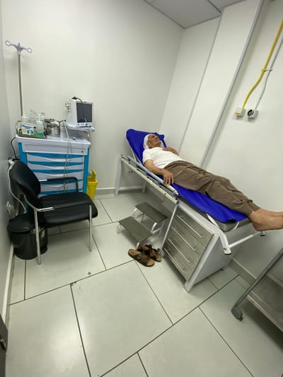 In a room within Baraki, Algeria, an elderly man lies on a blue medical examination table. He is wearing a white t-shirt and brown striped pants. His head is propped up on a pillow, and his right hand rests on his chest. To the left of the table is a set of metal steps leading up to it. Beside the steps, a black office chair is positioned. To the left of the chair, a dark gray trash can stands on the tiled floor. Behind the chair and to the left, a medical cart with blue drawers is visible, holding various medical supplies, including bottles and containers. A portable monitor with a screen is mounted on top of the cart. A drip stand is also present to the left of the cart. Yellow medical gas lines are visible running along the white wall to the right of the examination table. Brown sandals are placed on the floor near the steps. The room has white walls and a tiled floor.