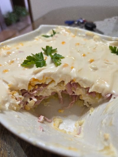 A white, rectangular serving dish, partially emptied, contains a layered food item. The surface of the food is covered with a creamy white sauce speckled with small orange-yellow particles and garnished with fresh green parsley sprigs. A section has been removed from the dish, exposing internal layers consisting of shredded pink ham, diced yellow and white material consistent with hard-boiled egg, and other light-colored components. The dish rests on a dark, textured surface. The background is out of focus, showing a blurred green plant in a pot to the left and a set of keys with a blue fob to the right. The scene is located in Negorci, North Macedonia.