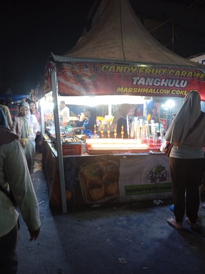 The image depicts a street food stall in what appears to be Kauman, Indonesia at night. The stall is selling "Candy Fruit Caramel Tanghulu" and "Marshmallow Coklat" as indicated on the banner. A woman wearing a headscarf is standing at the counter, apparently a customer. Another woman, wearing a headscarf, appears to be purchasing the item, with a worker standing behind the counter at the stall. A group of women can be seen to the left side of the stall. The stall is lit up, with skewers of the candy treats displayed. The stall front has an advertisement for "Crocs" and another ad that appears to say "sandal waffle." The ground is asphalt with bits of debris.