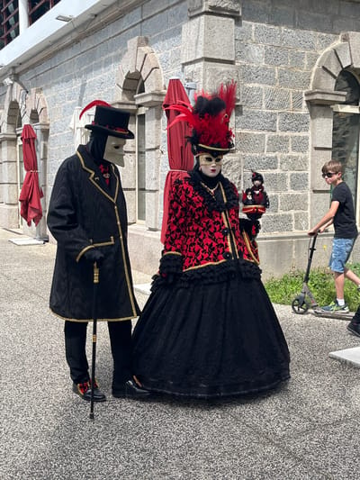 In Saint-Galmier, France, two individuals stand in ornate Venetian Carnival-style costumes. One wears a black top hat with a red feather, a black jacket with gold trim, black pants, black shoes, and a white mask. The other wears a red and black gown, black shoes, and a white mask with red feathers. Both appear to be wearing costumes related to a festival or event. A child on a scooter is in the background, near a stone wall. A gray stone pavement is visible. Parasols are visible in the background.