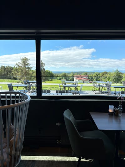 This image, taken from inside a dark room with a large window, offers a view of an outdoor seating area with tables and chairs, overlooking a landscape. The outdoor area features numerous tables and chairs arranged on a paved patio, separated from a grassy area by a glass railing. Beyond the patio, a building with a red roof is visible, surrounded by trees and rolling hills that extend to the horizon under a partly cloudy blue sky. The interior of the room shows a white crib in the foreground on the left, and a table with a lit candle and salt and pepper shakers on the right. This scene is likely at a restaurant or event venue in Östersund, Sweden, given the distinctive landscape and architecture that aligns with the region's characteristics. No people are visible in the image.