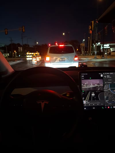 The image shows the interior of a Tesla vehicle at night, with the driver's perspective looking out at traffic. In the foreground, the steering wheel and a portion of the dashboard are visible. The central infotainment screen displays navigation, a camera feed, and media controls. Outside the car, traffic lights are red, indicating the vehicle is stopped. Construction barrels and a white SUV are visible in the road ahead. The sky is dark, suggesting it is nighttime. The location is identified as Buffalo Grove, United States of America.