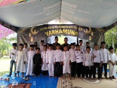A group portrait taken outdoors in Jambo Dalam, Indonesia, features twenty individuals beneath a temporary tent structure. Nineteen young males, primarily children or adolescents, stand in two rows facing the camera. Most wear white or light-colored traditional Islamic attire, including long shirts and various head coverings such as songkok or peci. One child on the far left wears a white shirt with jeans. Another child near the center wears a black long-sleeved garment. Two adult males stand towards the back-center, also in Islamic attire and headwear. All subjects appear to be posing, looking towards the camera.
The setting is under a white and patterned fabric canopy. A dark banner prominently displays "LEMBAGA PENDIDIKAN ISLAM YARHAMUNA" in yellow text, flanked by crescent moon and lantern motifs, across the rear of the tent. Other banners are partially visible and textually reversed on the upper tent fabric. The ground beneath the subjects consists of a blue tarp, with a decorative brown and cream patterned rug laid over it in the foreground where the subjects stand. Several plastic cups, bottles, and cables are scattered on the blue tarp to the left. Beyond the tent's perimeter, green foliage and trees are visible under an overcast sky.