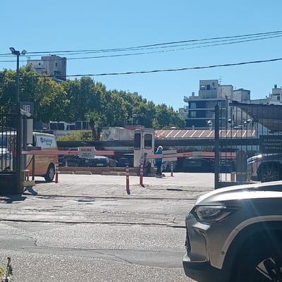 The image depicts the exit area of a commercial parking lot in Montevideo, Uruguay, under a clear blue sky. The immediate foreground on the right displays the front quarter of a silver/grey SUV. The paved lot surface is visible, showing cracks and irregular patches.

Central to the scene are two red and white striped barrier gates; the left gate is raised, indicating an open exit, while the right gate is lowered. Between these, a white security booth with a canopy is situated. Two individuals are present near this booth: one person with light-colored hair, dressed in a light blue top and dark pants, stands facing left; the second individual, wearing a light-colored shirt and dark pants, is seated or leaning against the booth, facing right.

A yellow "Parkings Group" commercial van is positioned at the left exit lane. Above the raised barrier, a sign reads "SALIDA" (Exit), with "FIESI MARKET" and additional text in Spanish visible below. A tall, slender pole to the left of the barrier displays the number "16" and appears to incorporate a camera or sensor. Orange traffic bollards are distributed across the paved area. Within the parking facility, several dark-colored parked vehicles are discernible. On the right, a black metal fence features a white sign in Spanish, stating, "EL ESTACIONAMIENTO PERMANECERÁ CERRADO DE 22:00 hs a 08:00 hs." (The parking lot will remain closed from 10:00 PM to 08:00 AM). The background consists of green-leafed trees and various urban buildings, some multi-story, under a cloudless sky traversed by utility lines.
