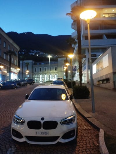 A white BMW hatchback is parked on a cobblestone street in Muralto, Switzerland, at dusk. The car has its license plate visible, reading "TI-36026". In the background, a modern building with balconies and outdoor lighting stands beside palm trees. Further in the distance, a street lamp illuminates a parking sign with the letter "P". The sky is a deep blue, transitioning into night, with faint lights visible on a distant hillside. Several other vehicles are parked on the street.