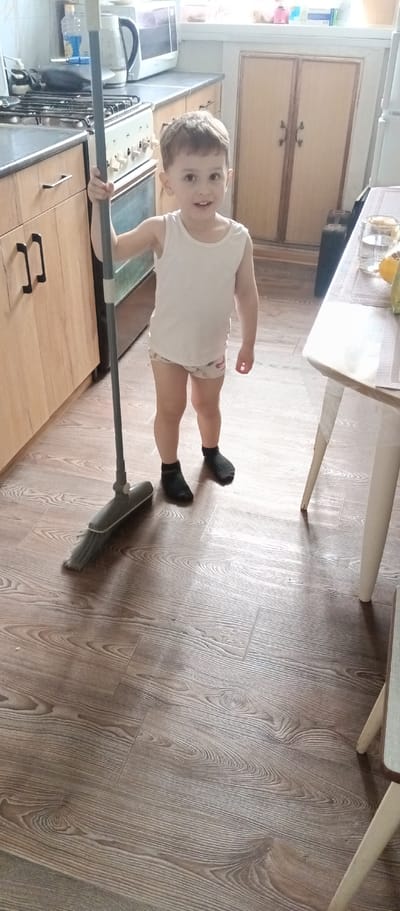 A young child stands in a kitchen in Irbit, Russia, holding a broom. The child is wearing a white tank top, patterned underwear, and black socks. The kitchen features wood-grain flooring, wooden cabinets, a stove, and a dining table with items on it. A broom leans against the cabinets, and the child is looking at the camera.