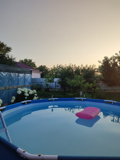This is a medium-shot, eye-level, vertical, outdoor photograph of an inflatable pool on a clear, calm day. The pool is filled with bright blue water, and a single pink inflatable mattress floats on its surface. The pool's blue and white frame is visible, along with a white ladder on the left side.

In the background, behind the pool, there is lush green foliage and trees. A light-colored house with a reddish-brown roof is partially visible behind the trees. A fence, appearing to be made of corrugated metal, runs across the middle ground. To the left, a structure covered in blue plastic sheeting and dark netting is also visible.

The sky is a pale, hazy orange, suggesting either early morning or late evening. The overall mood is peaceful and serene. The image was taken in Abinsk, Russia. There is no visible text in the image.