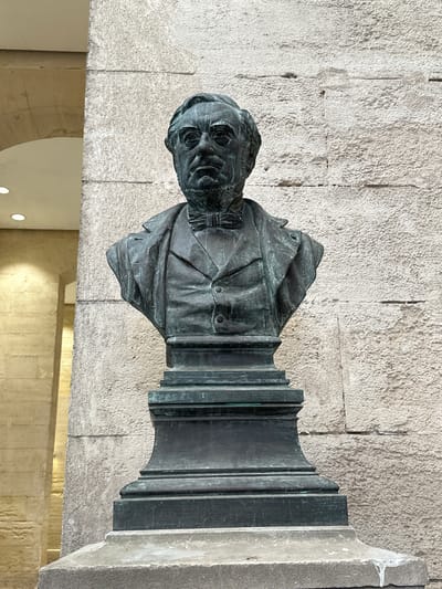 This is a medium-shot, eye-level, outdoor photograph of a bronze bust of a man. The bust is dark green and weathered, with visible details of the man's face, hair, and clothing. He has a prominent nose, deep-set eyes, and is wearing a suit with a bow tie. The bust is mounted on a dark, stepped pedestal, which in turn rests on a lighter stone base.

The background consists of a rough-textured, light-colored stone wall, likely limestone. To the left of the bust, there is a portion of an arched structure, suggesting an arcade or building facade, made of lighter-colored stone with visible horizontal lines. The overall lighting is subdued, with some illumination coming from the left.

The bust is located in the Place de l'Horloge in Nîmes, France, specifically in front of the Maison Carrée.