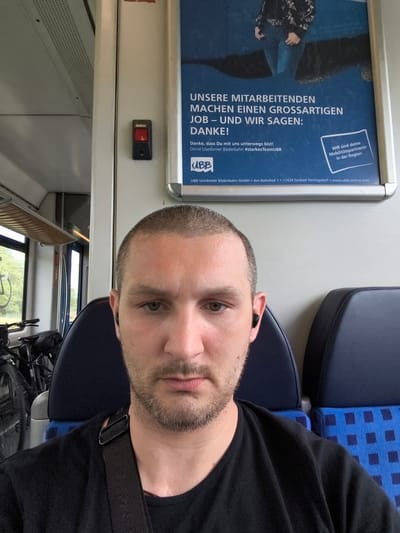 The image is a selfie taken by a man inside a train. He has a buzz cut and is wearing a black t-shirt and black wireless earbuds. He is looking directly at the camera with a neutral expression. Behind him, out of focus, are dark blue train seats, some with blue checkered upholstery. The man is wearing a black strap across his chest, possibly from a bag.

To the left of the man, a bicycle is visible, suggesting a mode of transport for passengers on this train. Further to the left, a window shows a glimpse of greenery outside, indicating it might be daytime.

On the wall behind the man, a large poster with blue and white text is displayed. The text is in German and appears to be an advertisement or a message from the train company, "UBB Usedomer Bäderbahn". Some of the visible text includes "UNSERE MITARBEITENDEN MACHEN EINEN GROSSARTIGEN JOB – UND WIR SAGEN: DANKE!" which translates to "OUR EMPLOYEES MAKE A GREAT JOB – AND WE SAY: THANK YOU!". The poster also includes the company logo and website.

Near the poster, a red "STOP" button is mounted on the wall. The overall scene suggests a journey on a local train in Germany, likely during daylight.