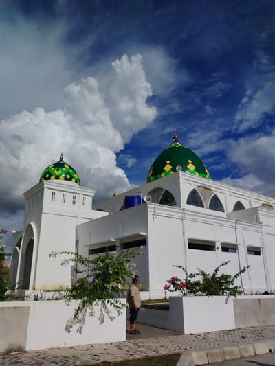 The image depicts a mosque with a prominent green and yellow checkered dome, set against a dramatic sky filled with large, white cumulus clouds and patches of deep blue. The mosque is a modern white structure with arched windows and a smaller dome visible on the right. In the foreground, a man in a light-colored shirt and dark pants stands facing away from the camera, looking towards the mosque. He is standing next to a white planter box filled with flowering plants, some with white blooms and others with red. The ground in front of the planter box is paved with bricks. The image is likely taken in the city of Keureuweungkrueng, Indonesia, based on the provided context, though this is not visually verifiable from the image itself. The overall scene conveys a sense of tranquility under a dynamic sky.