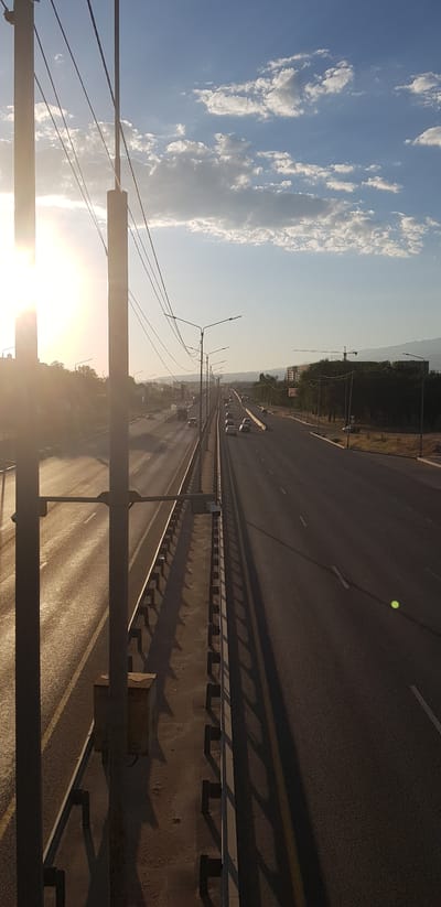 The image depicts a highway with multiple lanes, a pedestrian walkway, and overhead power lines, likely in or near Іргелі ауылдық округі, Kazakhstan. The sun is low in the sky, casting long shadows and creating a lens flare on the left side of the image. There is moderate traffic on the highway, with several cars visible. Streetlights line the highway, and buildings and trees are visible in the background, along with distant hills or mountains. A construction crane is also visible in the distance. The scene appears to be a typical urban or suburban highway environment during sunset.