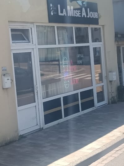 The image displays the street-level facade of a commercial establishment in Wolfisheim, France. The central subject is a shopfront with a light yellow building exterior. A prominent black sign above the entrance reads "LA MISE A JOUR" in white capital letters, accompanied by a white graphic resembling a barber's pole or razor.
The shopfront consists of a white-framed glass door on the left and multiple large, white-framed glass display windows to the right. A small, white-framed casement window positioned above the main door is partially open outwards. Inside the primary display window, a vertical neon sign shaped like a barber's pole emits a distinct blue and pink light. Reflections on the glass panes show a paved road with white pedestrian crossing stripes and a dark-colored vehicle. Faint interior signage, including text such as "COIFFEUR" and "DEPOT!", is visible through these reflections. The lower sections of the display windows are opaque, featuring dark panels with a single horizontal gold stripe.
A light-grey tiled sidewalk occupies the foreground, exhibiting a sharp diagonal shadow consistent with direct sunlight. A white rectangular utility box or mailbox is mounted on the building's facade to the immediate left of the shop's door. A similar, though partially obscured, white box is visible on the adjacent building to the right. Small patches of green vegetation are present at the base of the wall on the right side of the shop. No individuals are visible, and the scene appears static.