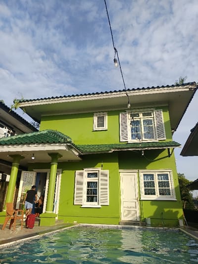 The image depicts a two-story lime green residence with a swimming pool in the foreground, located in Palasari, Indonesia. The building features a dark green tiled roof with white trim and minor visible plant growth. White-framed windows with decorative grilles are present on both floors, some equipped with white louvered shutters. A covered porch on the left side of the ground floor is supported by two lime green columns.

In the mid-ground, on the porch, a person, possibly male, wearing a dark t-shirt and light shorts, stands pulling a red rolling suitcase. Another indistinct individual is partially visible behind them, near the open doorway. A light brown wicker-style chair is positioned on the left side of the porch. A string of exposed bulb lights is strung horizontally across the upper portion of the frame, above the second-story windows. The swimming pool, filled with clear, rippling blue-green water, occupies the lower third of the frame directly in front of the house. The sky above is blue with scattered white clouds.
