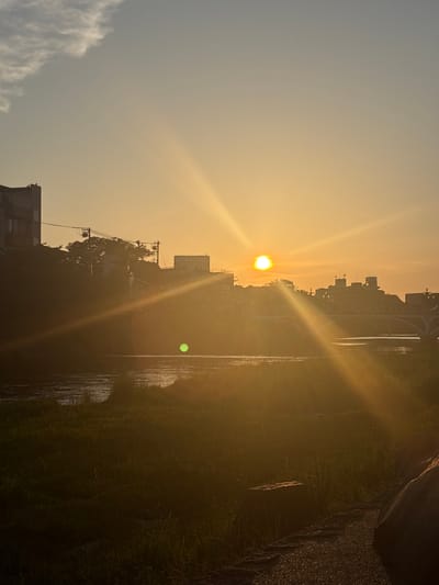 This image captures a sunset or sunrise over an urban river in Kanazawa, Japan. The sun, a brilliant white-yellow orb, is positioned in the upper center-right, emitting intense golden rays and lens flares that traverse the scene diagonally. The sky transitions from a pale blue-grey with diffuse clouds in the upper left to a warm, vibrant golden-orange closer to the horizon and the sun.
The river, characteristic of urban waterways in Kanazawa such as the Saigawa or Asanogawa, flows across the mid-ground, displaying ripples on its surface and reflecting the warm ambient light. The river's banks are lined with dark, silhouetted vegetation. On the left bank, dense trees and bushes obscure a hillside, behind which the outlines of multi-story buildings and utility infrastructure, including power lines, are visible.
The right bank features a silhouette of urban development, including multiple buildings forming a skyline. A prominent multi-arch bridge spans the river towards the right side of the frame. In the foreground on the right, a dark grassy bank is visible, interspersed with large rocks and a gravel path. Individual blades of grass are discernible, some possibly catching dew or moisture. A distinct, circular green lens flare appears in the lower-mid section of the image, slightly to the left of the sun's central light path. The overall lighting conditions suggest either early morning or late evening, bathing the landscape in a warm, hazy glow.