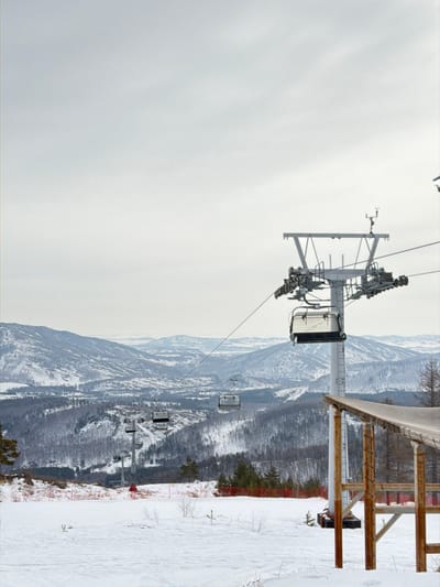 The image displays a winter scene at a ski resort in Novoabzakovo, Russia. An overcast, light grey sky occupies the upper portion, transitioning to a snow-covered mountainous landscape below. In the mid-ground on the right, a prominent metal ski lift pylon stands, supporting an array of cables, pulleys, and a weather instrument at its apex. An enclosed white ski gondola is suspended from the cables near this pylon. Two additional white ski gondolas are visible further down the slope to the left, appearing to descend. Another ski lift pylon is present in the mid-distance, supporting the cables.

The foreground consists of a gently sloping, snow-covered terrain. To the lower right, a wooden structure, likely a loading platform for the ski lift, features a sloping, covered roof and exposed wooden supports. In the mid-ground, to the left of the central pylon, an individual wearing red attire is standing on the snow, positioned near a stretch of orange safety netting. The background reveals a vast panorama of undulating, snow-dusted mountains and valleys, densely covered with a mix of bare deciduous and evergreen trees extending to the horizon.