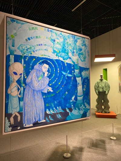 The image depicts an art exhibition featuring a large, vibrant painting on the left and a sculpture on the right. The painting is a surreal composition with a dominant blue palette, featuring two figures: an alien with large eyes standing to the left, and a person in a blue robe with a golden pattern in the center. Both figures are depicted in profile, with the robed figure appearing to be speaking or gesturing. The painting is adorned with numerous Chinese characters and some English text, interspersed with stylized trees and birds. The overall style suggests a blend of traditional and modern artistic influences.
To the right of the painting, a sculpture of a figure made of stacked, green, textured elements stands on an orange pedestal. The sculpture has a metallic, faceless head and resembles a layered, organic form. The exhibition space has a dark ceiling with exposed beams and lighting fixtures, and the walls are a neutral, light color. White ropes and small stands are present in the foreground, delineating the viewing area and casting shadows on the floor. The scene appears to be indoors, within a museum or gallery setting. The location is identified as Jinniu District, China.