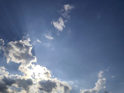 The image shows a sky with clouds and sun rays over Mykulychyn, Ukraine. The sky is blue with scattered white and gray clouds. The sun is partially obscured by clouds, creating dramatic sunbeams. The overall impression is one of a bright, possibly partly cloudy day.