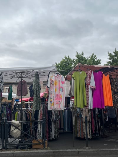 The image depicts an outdoor clothing market in Villeparisis, France, under a cloudy sky. Several stalls are visible, with racks of clothing hung on display. The foreground shows a metal railing with a white bag and some black trash bags. Behind the railing, a variety of dresses and other garments are arranged on hangers and racks. Some of the clothing is brightly colored and patterned, while other items are more muted. Several price tags are visible, including one for "5 €" and another for "10 €". In the background, trees and buildings can be seen, as well as a few people browsing the stalls. One person, wearing a blue shirt and dark shorts, is partially visible on the right side of the image, facing away from the camera. Another person, a woman with blonde hair, is visible behind the racks of clothing on the left. The market appears to be set up on a paved surface, likely a street or square.