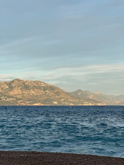 The image shows a tranquil seascape with a pebble beach in the foreground, a calm blue sea in the middle ground, and a majestic mountain range in the background. The sky above is a soft gradient of light blue and white, with subtle cloud formations. The mountains appear to be partially populated with scattered white buildings, suggesting a coastal settlement. The overall impression is one of natural beauty and serenity, likely captured in the early morning or late afternoon given the soft lighting. The location is identified as Lastva Grbaljska, Montenegro. There are no visible people or specific actions depicted.