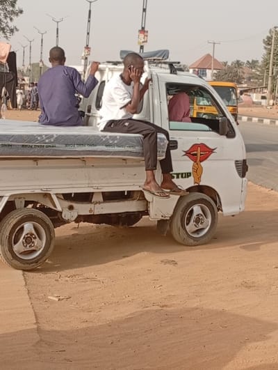 The image depicts an outdoor scene in what appears to be a dry and sunny environment, characteristic of Kabuga, Nigeria. A white pickup truck, possibly a light commercial vehicle, is the central focus, positioned on a dusty, reddish-brown road.

Two men are riding in the open bed of the truck. On the left, a man in a dark blue traditional garment is seated on a mattress, facing away from the camera, with his right arm raised in a gesture. To his right, another man, wearing a white t-shirt and dark pants, sits on the edge of the truck bed, his bare feet with flip-flops dangling. He is holding a smartphone to his ear, appearing to be on a call. A white mattress, bearing the repeated text "HENG FENG FOAM," covers the truck bed.

Inside the truck's cabin, a person wearing a maroon or reddish-brown headscarf is visible. A notable detail on the truck's passenger side is a sticker featuring red lips with a finger shushing them, and the partially visible word "STOP" beneath. The truck itself appears to be used, showing some wear.

In the background, other vehicles, including a yellow tricycle, are partially visible. Various buildings and utility poles line the road, suggesting an urban or semi-urban setting. The bright daylight and clear sky indicate a sunny day. The scene captures an everyday moment of transportation and activity.