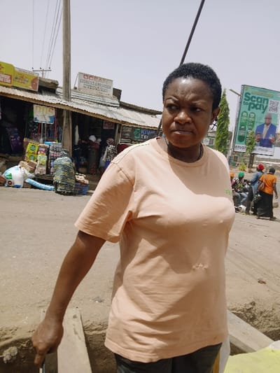 The image portrays an outdoor urban street scene in Kadoma, Zimbabwe, under bright daylight. A dark-skinned woman occupies the central foreground, positioned from the waist up, facing slightly towards the viewer's right. She exhibits a serious expression with furrowed brows. Her hair is short, dark, and curly. She wears a plain, light peach-colored short-sleeved t-shirt and dark trousers. Her left arm is extended downwards, and her left hand grasps a dark object positioned near a concrete edge or barrier. A small, dark, cross-shaped earring is visible in her left ear.

Behind the woman, a commercial area features multiple single-story buildings with corrugated metal roofs. A prominent sign on one building reads "OCEAN COSMETICS & PROVISION," with another partially visible sign below it indicating "KWI TEI." Various packaged goods and merchandise are displayed outside these shops. Several individuals, primarily women dressed in patterned fabrics, are discernible near the storefronts. The ground surface is unpaved dirt or sand.

To the right in the background, a large vertical billboard is visible, featuring the text "Scan to pay" and an image of a man in a suit. A phone number beginning "07007" is partially displayed at the bottom of the billboard. Further in the background, additional individuals are observed, some standing, others appearing to be near a motorcycle or scooter. A utility pole extends vertically through the midground. The sky above is clear and bright.