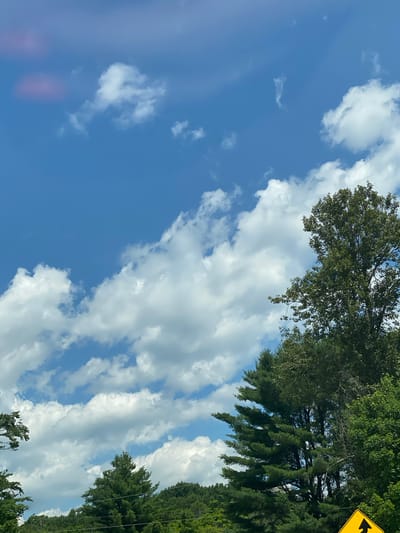 The image is a medium-angle shot looking upwards at a bright blue sky filled with scattered white cumulus clouds. The tops of several large green trees are visible along the bottom and right edges of the frame, with a prominent large deciduous tree dominating the right side. The scene appears to be set in a natural environment, likely outdoors during daylight hours. There is a yellow traffic sign with a black arrow pointing upwards in the bottom right corner, indicating a road or direction. The location is identified as Lac-Simon, Canada.