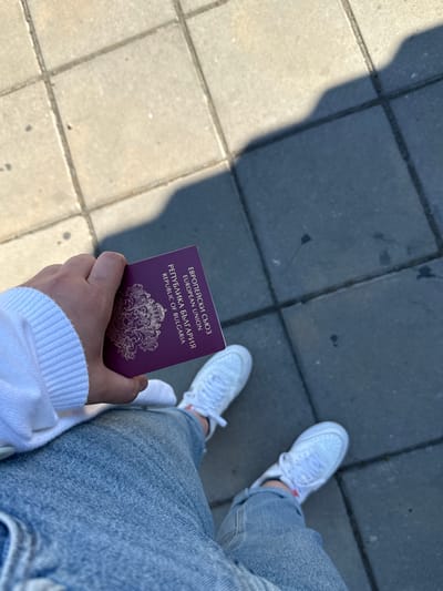 A person is holding a Bulgarian passport in their left hand. The passport is dark red with gold lettering and an emblem. The person is wearing a white hooded sweatshirt and blue jeans, and white sneakers. The background is a paved walkway with square concrete slabs. A shadow is cast across the walkway from the right side of the image, suggesting an overhead light source, possibly the sun. The scene appears to be outdoors in an urban or public area. The specific location is the Kapitan Andreevo border crossing point in Bulgaria.