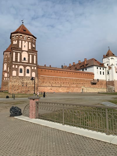 The image displays the Mir Castle Complex in Мірскі сельскі Савет, Belarus, under an overcast sky. The foreground features grey interlocking pavers and a low, dark-grey metal fence with circular decorative elements, supported by a short brick pillar on the left. Adjacent to the pillar, a dark, wheel-like decorative metal object is positioned on the pavers. Beyond the fence, a patch of sparse, dry grass is visible, with a set of grey steps ascending on the far right.
The Mir Castle dominates the mid-ground and background. On the left, a tall, multi-story tower with a red-tiled conical roof and a finial is prominent. This tower is constructed primarily of reddish-brown brick, accented with lighter pink plaster panels and white trim around its various arched and rectangular windows. Connecting this tower to the central part of the complex is a robust, red brick wall built upon a stone foundation. A black streetlamp stands on the paved area in front of this wall.
Further to the right, behind the wall, a white-faced section of the castle palace is visible, featuring multiple dark windows and red-tiled roofs with chimneys. A smaller, white-faced tower with a red-tiled conical roof and finial completes the visible structures on the right.
Several individuals are present on the large paved plaza area in front of the castle wall. One person in a light pink jacket and another in dark clothing are walking towards the left-center of the frame. Other figures are faintly visible further back. No explicit interactions between individuals are discernible.