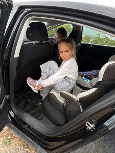 A young girl sits in the back seat of a black car, putting on a pink and silver shoe. A car seat is partially visible to her right. An adult with glasses is in the front seat. The car is parked outdoors, possibly in a rural or semi-rural area. The location is Provodina, Montenegro.