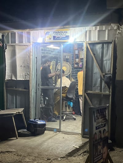 A dimly lit barbershop with a partially open front. Inside, a barber in black attends to a customer in a yellow patterned shirt. A fan circulates the air. An advertisement sign is visible on the right with images of haircuts. Another sign on the left reads "FAROUQ TAFEEDA". The shop appears to be simply constructed, with a metal frame and various items stacked along the exterior. A blue generator sits on the ground to the right.